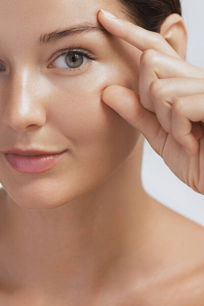 Крупный план молодой женщины, нежно прикасающейся к коже вокруг глаза, демонстрирующей свой сияющий цвет лица. Этот образ идеально подходит для рекламы красоты, анти-старения и скинкаре, фокусирующейся на деликатной
