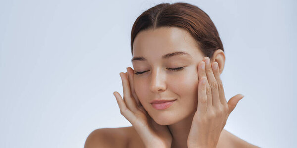 Расслабленная женщина с светящейся кожей нежно касается своего лица обеими руками, глаза закрыты в мирном выражении. Идеально подходит для косметических, лыжных и оздоровительных кампаний, пропагандирующих самоуход, гидратацию и