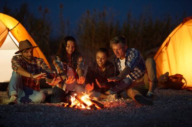 Bir grup arkadaş, kumsaldaki kamp yerlerinde turuncu çadırlarıyla kamp ateşi etrafında oturmuş serin bir gecede sıcaklığın ve arkadaşlığın tadını çıkarıyorlar..