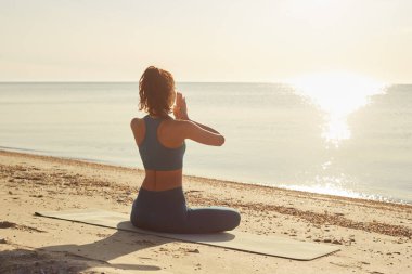 Bir kadın gün doğumunda plajda yoga yapıyor, okyanusa bakıyor ve o anın huzurunu kucaklıyor. Sağlık, farkındalık ve sahil kenarı rahatlama faaliyetleri için idealdir.. 