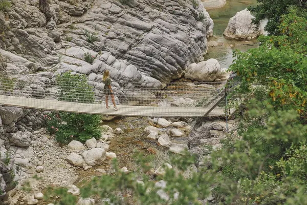 Bir kadın, Goynuk Kanyonu 'ndaki kayalık bir derenin üzerindeki asma köprüden geçiyor. Etrafı engebeli doğa ile çevrili. Çevre turizmi, yürüyüş ve açık hava macera deneyimleri için ideal.. 