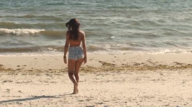 Kot şortlu ve bikinili genç bir kadın kumsalda denize doğru yalınayak yürüyor. Yaz için ideal, sahil yaşam tarzı, ve reklamlar veya seyahat tanıtımları için tatil kavramları. 