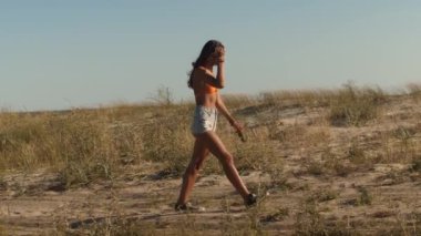 Turuncu bikinili ve kot şortlu genç bir kadın kumlu bir sahilde okyanusa doğru yürüyor. Yaz yaşam tarzı, seyahat ya da plaj tatili tanıtım içeriği için harika. 