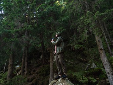 Bir fotoğrafçı elinde kamerayla bir kayanın üzerinde duruyor ve sık bir ormanın güzelliğini yakalıyor. Doğa fotoğrafçılığı, macera ve vahşi doğanın cazibesi için mükemmel bir tema.. 