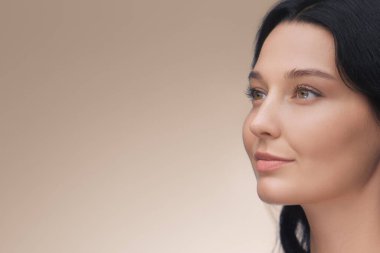 Koyu renk saçlı, parlak tenli, bej bir arka plana dik dik bakan zarif bir kadının yakın plan profili. Cilt bakımı, sağlık ve güzellik reklamcılığı kampanyaları için ideal.