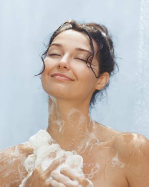 Saçları topuz şeklinde olan sakin bir kadın, göğsüne yakın bir lif tutan köpüklü bir duşun keyfini çıkarırken mutlu bir şekilde gülümsüyor. Bu görüntü, saf rahatlama ve kendine bakmanın bir anını yakalar.