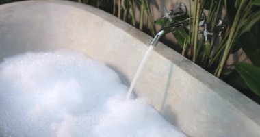 Lüks bir açık hava taş küveti, yemyeşil bitkilerle dolu tropik bir ortamda köpük ve akan suyla dolduruluyor. Yüksek kaliteli spa, rahatlama ve doğa temalı kavramlar için idealdir.