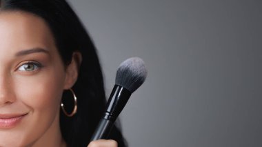 Makyaj fırçası tutan, parıldayan derisini ve şık halka küpelerini gösteren yarım yüzlü bir kadın portresi. Güzellik, cilt bakımı ve makyaj uygulamasına odaklanmış kozmetik promosyonları için ideal