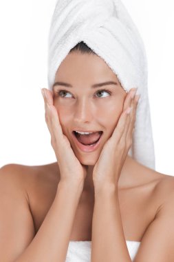 Kafasına havlu sarılmış heyecanlı bir kadın, cilt bakımı sırasında ellerini yüzüne dayayarak şaşkınlık ve neşe gösteriyor. Güzellik, özgüven ve oyunculuk temaları için ideal.