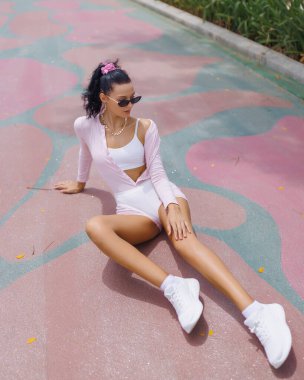 A confident woman in pink activewear sits on a colorful pavement, adjusting her sunglasses in a casual yet stylish pose. Her sporty look and vibrant surroundings highlight fitness, relaxation, and clipart