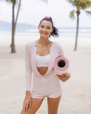 Tropikal bir plajda pembe yoga minderi tutan günlük spor kıyafetleri içinde gülümseyen genç bir kadın. Fitness, sağlıklı yaşam tarzı, sağlık ve tatil kavramları için idealdir. Yüksek kaliteli stok fotoğrafı..