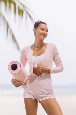 Genç bir kadın, tropik bir plajda pembe bir yoga minderi tutarken, günlük kıyafetlerle gülümsüyor. Sağlık, zindelik ve sağlıklı yaşam tarzı temaları için mükemmel. Tatil ya da dinlenme için ideal