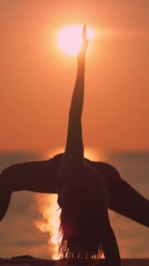 Bir kadın silueti, gün batımında kumlu bir plajda, esneklik, denge ve sakinliği ön plana çıkaran uzun bir kenar açı yoga pozu verirken yakalanır. Sağlık, yoga ve zindelik için ideal.