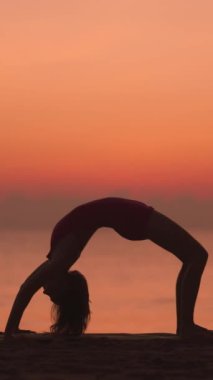 Bir kadının dikey videosu gün doğumunda plajda hareketli gökyüzüne karşı siluet şeklinde poz veriyor. Bu dingin yoga sahnesi esnekliği, gücü ve farkındalığı vurguluyor, sağlık için ideal.