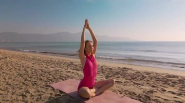 Bir kadın, güneşin doğuşunda, sakin okyanusa bakan kumlu bir sahilde, nilüferde meditasyon yapıyor. Farkındalık, sağlık, açık hava yogası ve doğada huzurlu meditasyon kavramları için mükemmel..