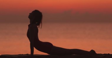 Bir kadın gün batımında kumlu bir plajda yoga yapıyor. Kobra gibi poz veriyor, aşağı doğru köpek gibi ve bacak kaldırıyor. Farkındalık, esneklik ve sakin doğa yogası için mükemmel..