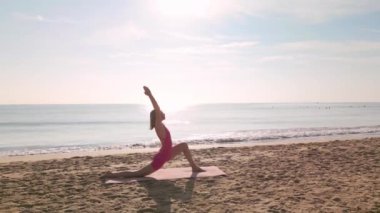 Pembe kıyafetli bir kadının, güneşin doğuşunda kumlu bir sahilde yoga yaptığı, okyanusa bakan sakin bir sahne. Farkındalık, rahatlama, fitness ve plaj yaşam tarzı kavramlarını desteklemek için mükemmel..