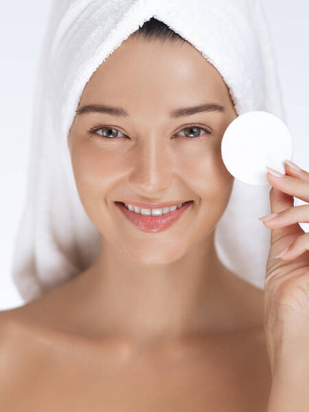 Улыбающаяся женщина с светящейся кожей и завернутыми в полотенце волосами держит хлопковую подушку, как часть своей рутины. Идеально подходит для продвижения красоты, хорошего самочувствия и кожи, делая акцент на здоровой, сияющей коже.