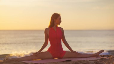 Pembe aktif giysili bir kadın gün batımında plajdaki bir minderde yoga yapıyor. Huzurlu okyanus ve altın gün batımı, refahı, esnekliği ve esnekliği desteklemek için ideal barışçıl bir atmosfer yaratır.