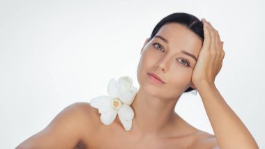 Çıplak omuzlu zarif bir kadın ve boynuna yakın duran beyaz bir nilüfer çiçeği, başını nazikçe elleriyle destekliyor. Minimalist ortam ve yumuşak ışıklandırma güzelliği ve rahatlığı vurguluyor.