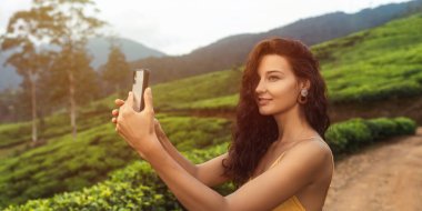 Kıvırcık saçlı genç bir kadın, dağ manzaralı, gür bir çay tarlasında akıllı telefonuyla fotoğraf çekiyor. Seyahat, doğa ve modern teknoloji temaları için ideal.. 