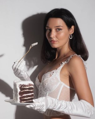 Beyaz iç çamaşırlı ve eldivenli baştan çıkarıcı bir kadın bir dilim pasta ve çatalla bir tabak tutuyor. Bu kaliteli stok fotoğrafı lüks, zarafet ve cazibeyi teşvik etmek için mükemmel.