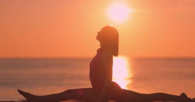 Pembe aktif giysili bir kadın gün batımında sicim yoga pozu veriyor. Huzurlu okyanus ve parlayan gökyüzü dingin bir ortam yaratıyor, fitness, farkındalık için mükemmel ve