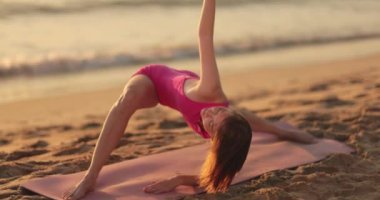 Bir kadın gün batımında kumlu bir plajda, bir kolu yukarı doğru uzanan köprü pozunun bir varyasyonunu sergiliyor. Esneklik, farkındalık, açık hava fitness ve dingin yoga pratiği temaları için ideal.