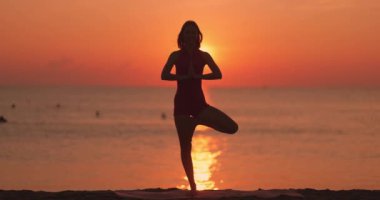Bir kadın gün batımında okyanusun üzerinde güneşin battığı bir plajda ağaç pozu veriyor. Fitness, farkındalık ve barışçıl doğa içeriği için ideal..