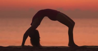 Bir kadın tek bacaklı köprü pozunu gün batımında kumlu bir plajda, arkada okyanusla birlikte veriyor. Farkındalık, esneklik, sağIık ve açık hava sporları konuları için mükemmel..