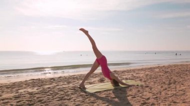 Bir kadın, gün doğumunda kumlu bir plajda sabah yoga seansı sırasında bir bacağı havada aşağı doğru köpek pozu veriyor. Esneklik, farkındalık, sağlık ve açık hava fitness temaları için idealdir..