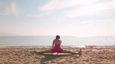 Bir kadın güneş doğarken, okyanusa bakan kumlu bir plajda yoga yapıyor. Huzurlu sahne, sakin ve doğal ortamda rahatlamayı, farkındalığı, zindeliği ve sağlığı teşvik eder..