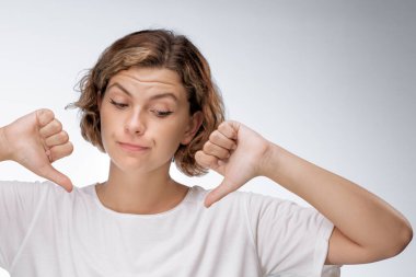 A woman in a white t-shirt gives a thumbs down with a displeased expression, indicating dissatisfaction. Ideal for advertisements related to customer feedback, product reviews, and dissatisfaction clipart