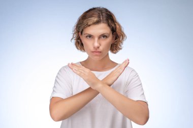 Beyaz tişörtlü, kollarıyla açık mavi arka planda X işareti yapan ciddi bir genç kadın. Reddetme, reddetme ya da anlaşmazlık kavramı. Reklamlar için yüksek kaliteli stok fotoğrafı ve