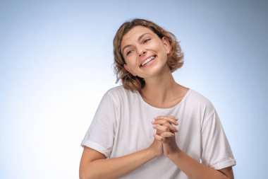 Beyaz tişörtlü neşeli genç bir kadın açık mavi arka planda elleri bağlı gülümsüyor. Reklam ve tanıtım materyallerinde mutluluk, pozitiflik ve neşe kavramları için idealdir.