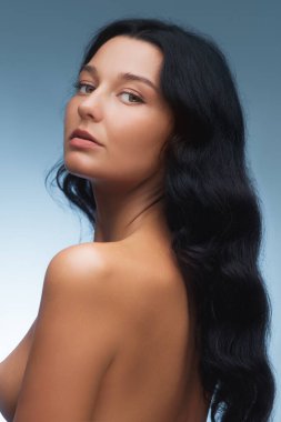 Uzun siyah saçlı, zarif, çıplak bir kadının portresi, mavi bir arka plana karşı sakin bir ifadeyle poz veriyor..