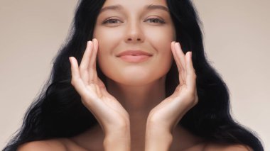 Gülümseyen Asyalı bir kadının simetrik yakın çekimi. Uzun siyah saçları ve pürüzsüz bir teni var. Elleriyle yüzünü çerçeveliyor. Güzellik, cilt bakımı ve kozmetik reklamları için ideal.