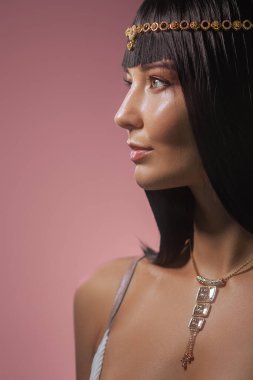 Kleopatra 'dan esinlenilmiş altın başlık ve kolyeyle süslenmiş, parlak siyah saçlı güzel bir kadın, pembe bir arka planda profil içinde yakalanır. Işıl ışıl cildi ve lüks havası