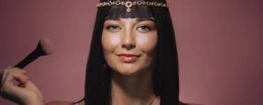 Kleopatra 'dan esinlenilmiş altın başlıklı şık bir kadın ve pembe arka planda makyaj fırçasını tutarken parlak siyah saçlarıyla gülümsüyor. Dengeli ifadesi ve lüks görünüşü...