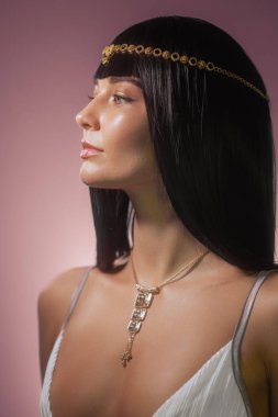 Kleopatra 'dan esinlenilmiş altın başlıklı ve parlak siyah saçlı şık bir kadın pembe arka planda poz veriyor. Işıl ışıl cildi ve zarif tavırları bu görüntüyü güzellik için ideal kılıyor.