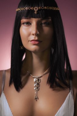 Parlak siyah saçlı, altın başlık ve kolye takan çekici bir kadın Kleopatra 'dan esinlenilmiş bir görünüme sahip. Durağan ifadesi ve gösterişli elbisesi, yumuşak ışıkla aydınlatılmış, bunu...