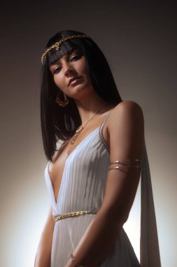 Pürüzsüz siyah saçlı, Kleopatra 'dan esinlenilmiş altın başlık ve mücevherlerle süslenmiş kendine güvenen bir kadın, zarif bir şekilde beyaz bir elbise içinde poz veriyor. Özgüvenli ifadesi ve lüks tarzı.