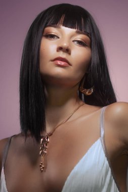 Parlak siyah saçlı, zarif makyajlı, altın takılarla süslenmiş, pembe eğimli arka planda poz veren çekici bir kadın. Üst düzey güzellik kampanyaları, lüks markalar için mükemmel.