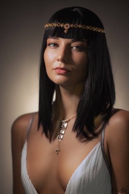 Parlak siyah saçlı, altın başlık ve kolyeyle süslenmiş güzel bir kadın narin bir ışıkta zarifçe poz veriyor. Dengeli ifadesi ve düşük kesimli beyaz bluzu lüksünü artırıyor.
