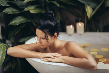 Young Asian woman enjoys a peaceful moment in a luxurious bathtub filled with citrus slices, surrounded by tropical plants and candlelight. Ideal for concepts of wellness, self-care, and spa retreats. clipart