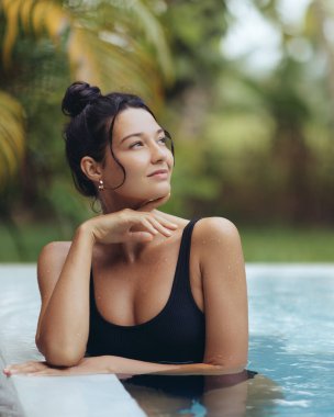 A young woman in a black swimsuit leans on the edge of a pool, enjoying a serene moment surrounded by tropical greenery. Perfect for luxury travel, wellness, and resort lifestyle concepts. clipart