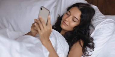 Esmer kadın, beyaz bir battaniyeye sarılıp, rahat bir şekilde yatakta uzanıyor ve akıllı telefonunu kullanıyor. Rahat ve modern bir yaşam tarzı yakalıyor, rahatlama, teknoloji ve yaşam tarzı temaları için mükemmel..