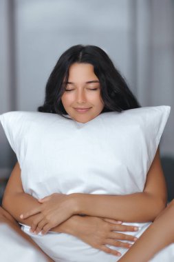 Huzurlu esmer kadın gözleri kapalı beyaz bir yastığa sarılıyor, sakinlik ve rahatlık saçıyor. Yumuşak doğal ışıklandırma, yaşam tarzı ve refah temaları için ideal, rahat ve huzurlu bir anı vurgular..