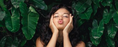 Kıvırcık saçlı, neşeli esmer kadın canlı yeşil tropikal yaprakların arasında poz verirken öpücük yolluyor. Güzellik, yaşam tarzı ve doğadan esinlenilmiş temalar veya sosyal medya içeriği için mükemmel.