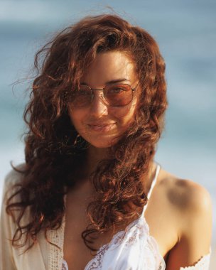 Dalgalı saçlı ve güneş gözlüklü şık bir kadın gün batımında plajda duruyor, akşamın sıcacık parıltısının tadını çıkarıyor. Moda, yaşam tarzı ve seyahat promosyonları için ideal.